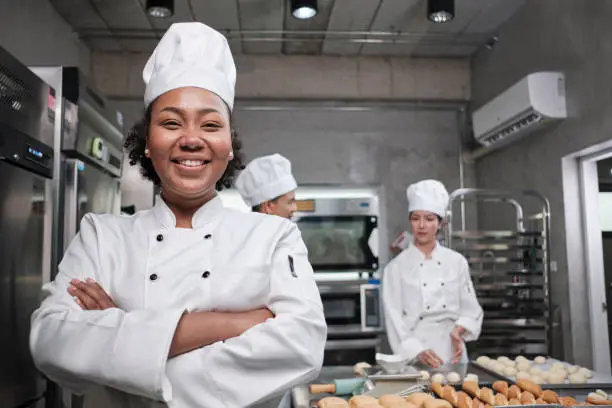 Young African American female chef in white cooking uniform looks at camera, arms crossed and cheerful smile with food professional occupation, commercial pastry culinary jobs in a restaurant kitchen.