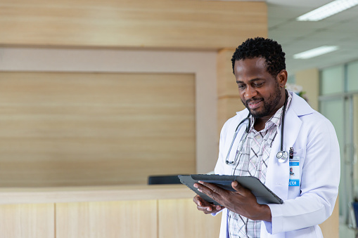 Male African doctor in white medical uniform fill patient medical history or anamnesis on tablet in hospital, man GP or physician consult client online on pad in modern clinic, healthcare concept