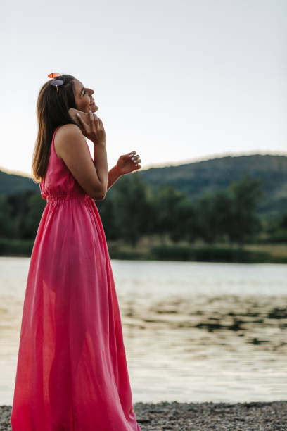 Beautiful young woman in dress talking on mobile phone by the lake at summer sunset