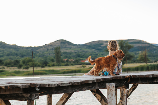 Young woman sitting with her dog on wooden pier and enjoying lake view on sunny summer day