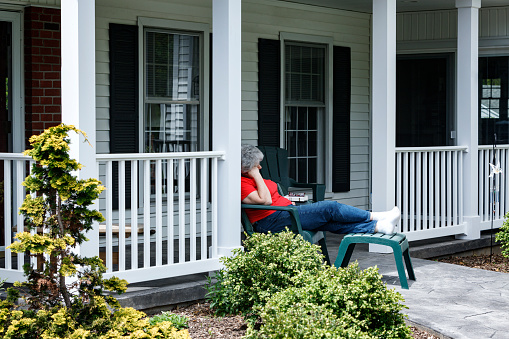 A relaxed senior adult woman reclining in a comfortable Adirondack chair had been reading a paperback book. She has put the book and her reading glasses down, and is sleeping peacefully with her head resting on her hand. She's snoozing on her front porch with her legs crossed at the ankles and her feet up on a footstool. She's wearing a bright red casual t-shirt, denim jeans, and white socks. A peek at a tranquil spring day at home in the suburbs near the city of Rochester, in western New York State.