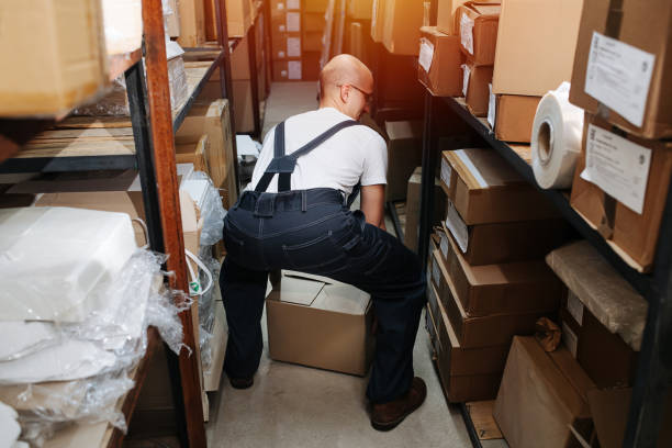vue arrière d’un employé d’entrepôt ramassant une boîte du sol en toute sécurité - giving file document stack photos et images de collection