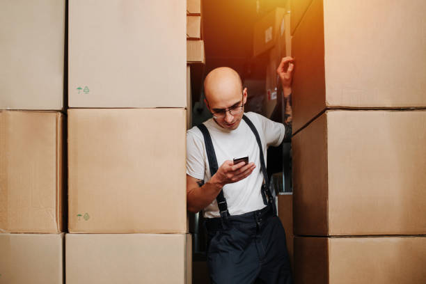 un ouvrier d’entrepôt audacieux et fatigué s’appuyant sur une pile de boîtes, regardant son téléphone - giving file document stack photos et images de collection