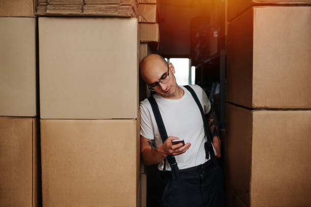 un employé d’entrepôt fatigué s’appuyant sur une pile de boîtes, regardant son téléphone - giving file document stack photos et images de collection