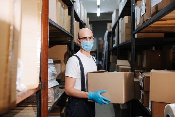 un employé d’entrepôt masqué tenant une boîte, les mains dans des gants bleus - giving file document stack photos et images de collection