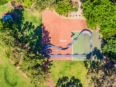 Aerial view looking directly down on portable BMX, Mountain Bicycle Pump Track on sports court in leafy public park. Plus basketball hoop, netball hoop, hopscotch marks.