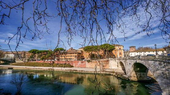 An idyllic and vibrant wide angle view of the Tiber Island and the Pons Cestius (Cestius Bridge), seen from the ancient Trastevere district, in the historic heart of Rome. Connected to the left and right banks of the Tiber river by two ancient Roman bridges, Pons Farbicius and Pons Cestius, the river island of Rome has always been associated with medicine and healing with the presence of the ancient temple of Aesculapius and subsequently of a hospital, still in function. In 1980 the historic center of Rome was declared a World Heritage Site by Unesco. Super wide angle image in 16:9 and high definition format.