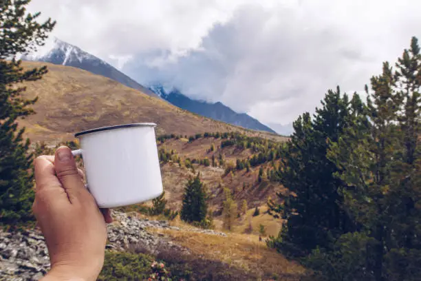Photo of Enamel white mug mockup with forest and mountains valley background. Trekking merchandise and camping geer marketing photo. Stock wildwood photo with white metal cup. Rustic scene, product template.