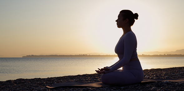 Healthy young woman sitting in yoga lotus pose with closing eyes in front of the sea, meditating at beach on sunrise.