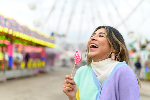 Portrait close up of happy young woman biting colorful lollipop in amusement park. Blurred background.