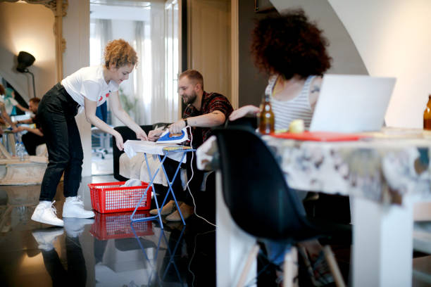 Young woman helping her male friend with the laundry, their flatmate working on laptop Young woman and man doing laundry at home and talking, their friend at the dining table working on laptop ad talking to them flatmate stock pictures, royalty-free photos & images