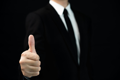 Unrecognizable smart businessman standing on the black background in photography studio and showing his thumb up to camera with copyspace. Satisfaction and excellent in business concept.