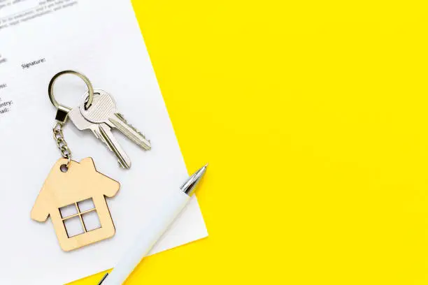 Keys with house shaped keychain and pen on real estate mortgage loan document, contract agreement to buy and construction new home, insurance, lease or rent apartments. Yellow background, copy space.