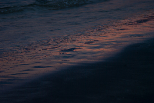 Dark pink surf on sea on sunset -  colorful pink, purple reflection of sunbeams and glare of sun in smooth deep blue waves and black line of beach, closeup, water texture, background.