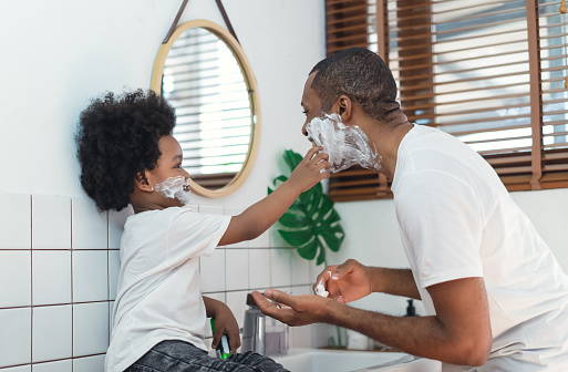 Happy African black father and little son with shaving foam on their faces looking at each other and laughing in the bathroom. Black family spend time together. Morning routine in bathroom concept.