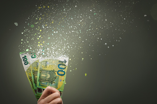 Several 100 Euro bills are dissolving into fine particles. Held by a hand in front of a neutral background with copy space.