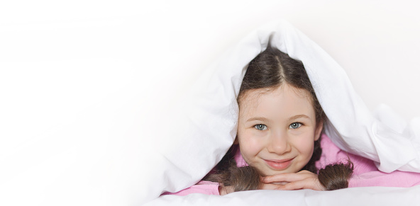 Portrait of a happy cute girl lying on white bed linen under a blanket. Copy space for text