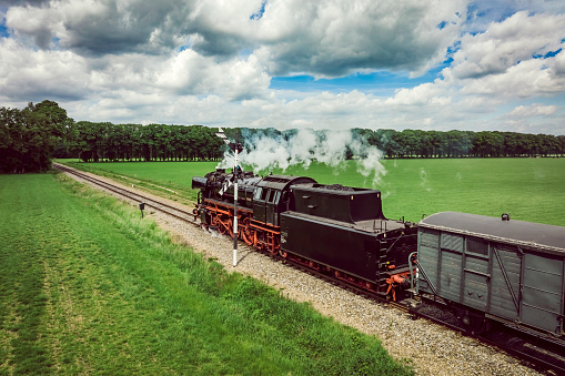 Steam train with smoke from the locomotive driving in the countryside with smoke coming from the chimney. The black and red locomotive is pulling passenger railroad car with tourists.