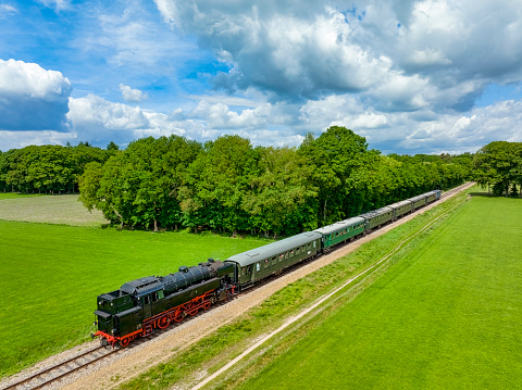 Steam train with smoke from the locomotive driving in the countryside with smoke coming from the chimney. The black and red locomotive is pulling passenger railroad car with tourists.