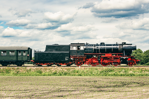 Steam train driving in the countryside with smoke coming from the chimney. The black and red locomotive is pulling passenger railroad car with tourists.