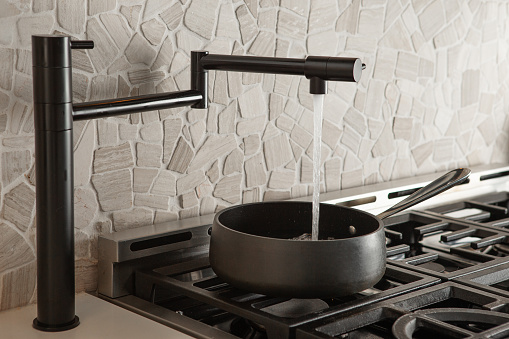 Black pot filler faucet filling up water in cooking pot on gas stove in kitchen with stone back splash
