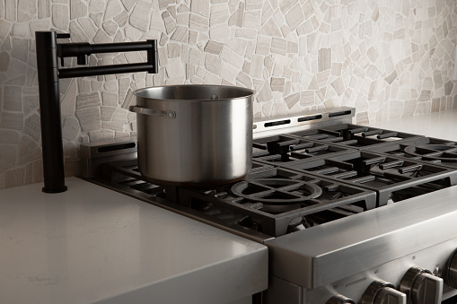 Black pot filler faucet over 6 burners gas stove in modern kitchen with stone back splash.