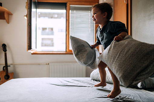 Joyful toddler jumping with pillow in his hands in bedroom