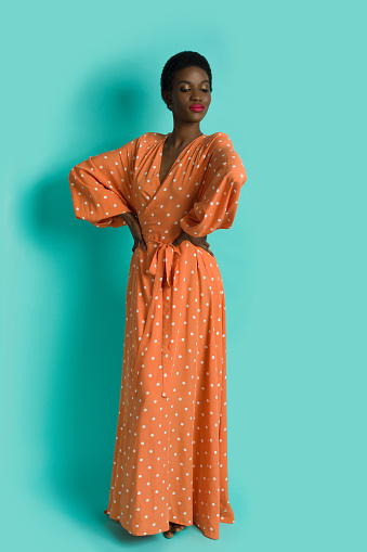 Young african woman wearing orange polka dot maxi dress and posing with hands on hip. Full length studio shot on turquoise background.