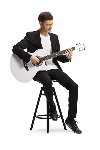 Elegant young man in a black suit sitting on a chair and playing a white acoustic guitar isolated on white background