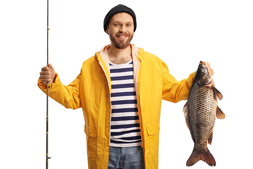 Happy young fisherman holding a carp fish and a fishing rod and smiling at camera isolated on white background