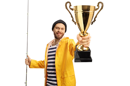 Happy young fisherman holding a fishing rod and showing a gold trophy cup isolated on white background