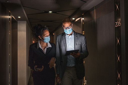 Hispanic business woman and mixed race business man wearing face masks, walking in a dark hotel corridor and using a digital tablet. Hispanic business woman holding her smartphone, while her business partner with eyegalesses showing to her some important working results on a digital tablet.