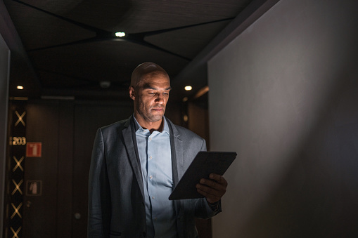 Portrait of serious mixed race business man in early 40s standing in a dark hotel corridor and using a digital tablet.