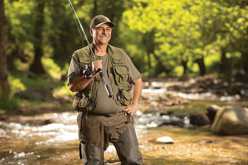Mature fisherman posing with a fishing rod next to a river