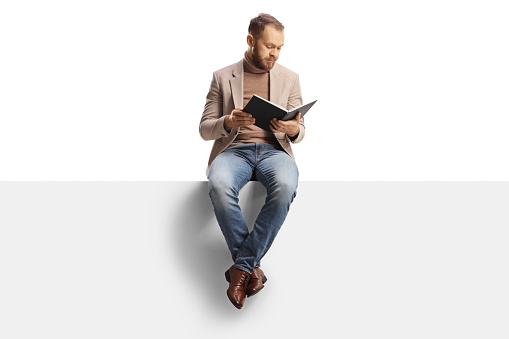 Young man reading a book and sitting on a blank panel isolated on white background