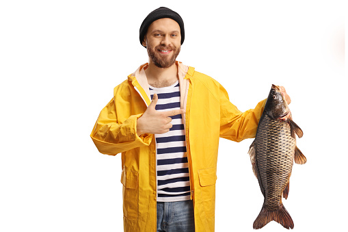 Young fisherman in a yellow raincoat holding a carp fish and pointing isolated on white background