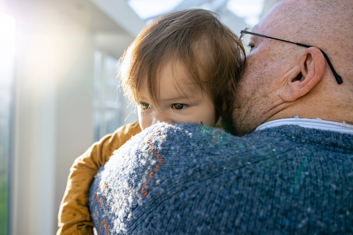 A close-up shot of a senior grandfather embracing and carrying his young grandson in his arms, he is resting his head on his grandfathers shoulder in their home in the north east of England.