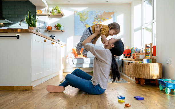 Mother's Baby Boy! A side-view shot of a mid-adult mother sitting on the floor of a living room with her young boy she is lifting him up in the air, they are wearing casual clothing playing and laughing together. toddlers playing stock pictures, royalty-free photos & images