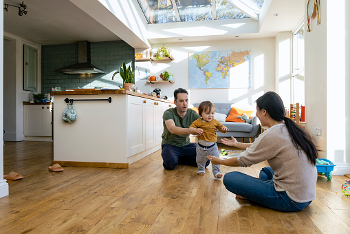 A side-view shot of a mid-adult mother and father sitting on the floor of a living room with their young boy who is learning to walk, they are wearing casual clothing and supporting their son taking his first steps.