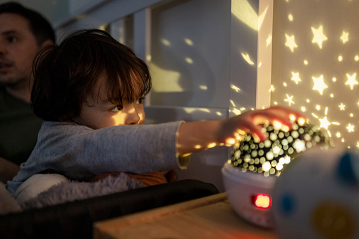 A close-up shot of a young boy wearing pyjamas lying in his bed with his father. He is looking at his new night light projector that is projecting stars and and the moon across his room with a cute smile.
