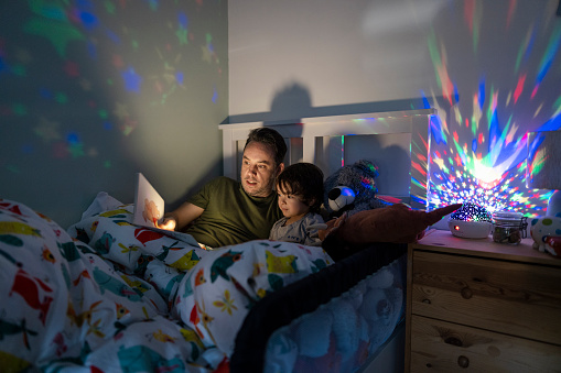 A wide-view shot of a young boy wearing pyjamas lying in his bed with his father. His father is reading him a bedtime story out of a book.  There is a colourful night light projector that is projecting stars across the boys room.