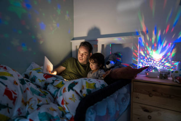 Father Reading Bedtime Story A wide-view shot of a young boy wearing pyjamas lying in his bed with his father. His father is reading him a bedtime story out of a book.  There is a colourful night light projector that is projecting stars across the boys room. bedtime stock pictures, royalty-free photos & images