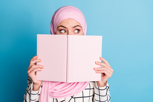 Close-up portrait of funny curious muslimah girl closing face book looking aside isolated over vivid blue color background.