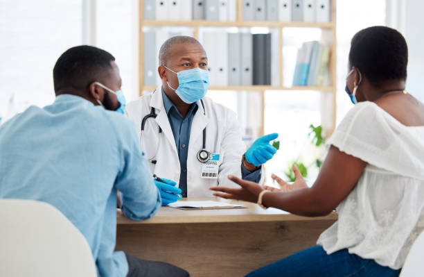 un couple afro-américain parle à un médecin du traitement de fertilité. médecin généraliste professionnel portant un masque pour se protéger du covid lors du contrôle. couple marié en consultation pour corona - husband wife family ethnic photos et images de collection