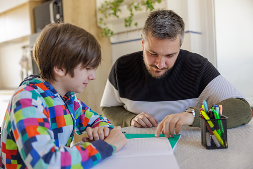 Father sitting with his elementary aged son at the table at home, helping him solve homework task
