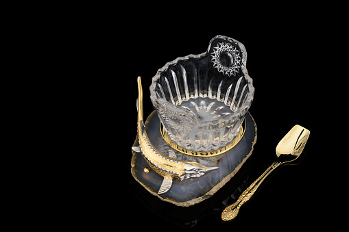 Gilded luxury crystal caviar bowl on stand made of natural quartz, carved brass fish figurine and dessert spoon on a black background.