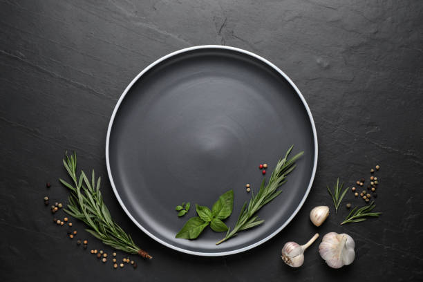 Dark plate with herbs, peppercorns and garlic on black table, flat lay