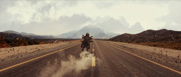 elegante motociclista brutale in sella a una moto in autostrada di velocità in autunno serale. sfondo cinematografico vintage e cielo nuvoloso drammatico. concetto di moda, stile e hobby - motorcycle biker sport city foto e immagini stock