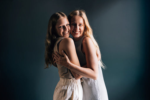 young beautiful brunette and blond haired girls hugs in front of gray background