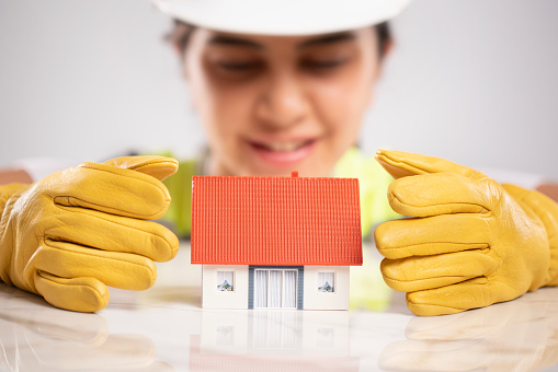 A model house is on the white background. A female engineer with yellow safety gloves and reflective vest. She is  looking at a model house and smiling.